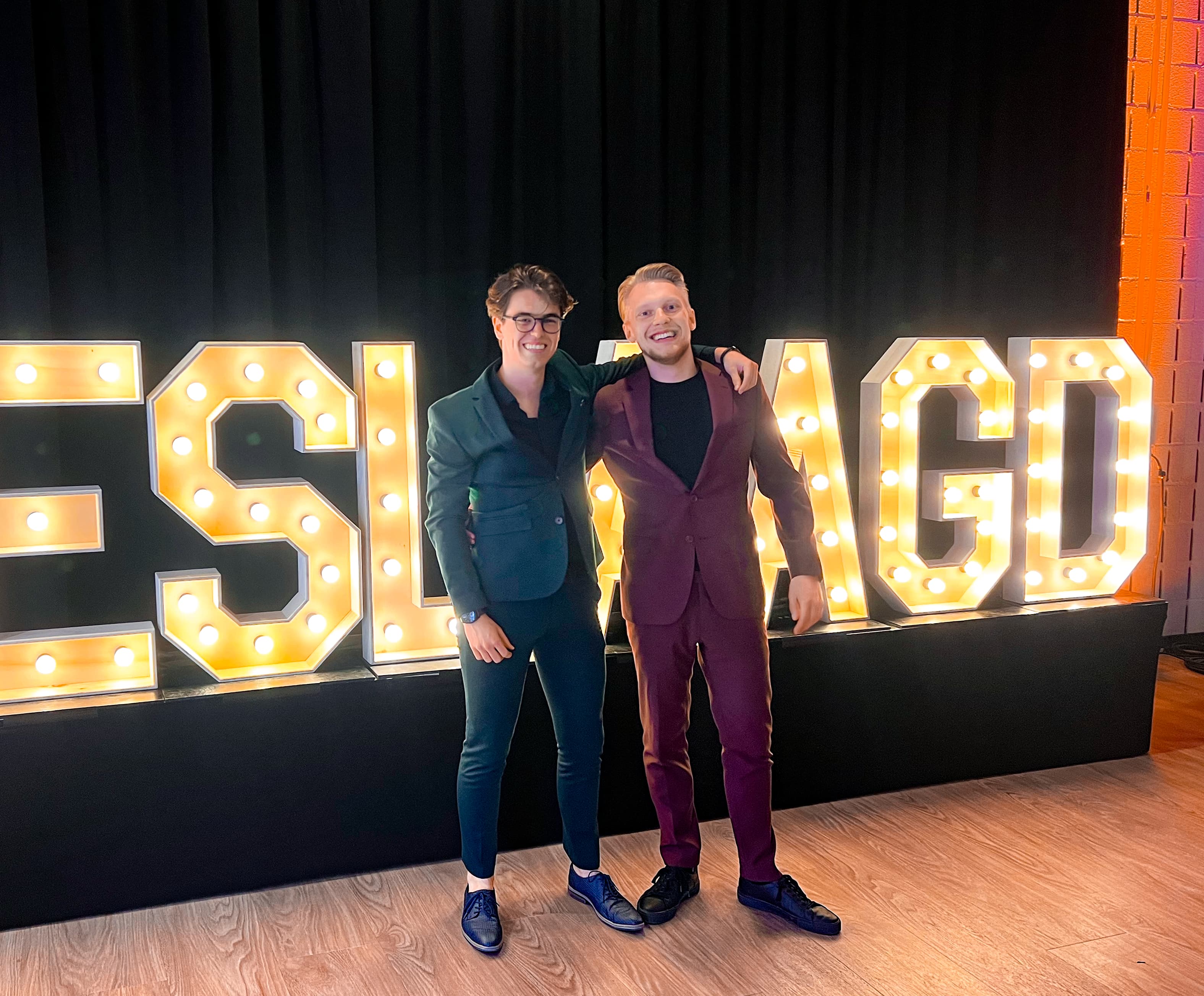 Rick en Rick poseren samen op de foto voor een groot bord met lichtletters 'geslaagd'. Rick Marcelissen heeft een rood pak aan en Rick van de Sande een groen pak.